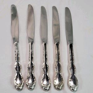 May include: Five silver butter knives with ornate handles. The knives have a curved blade and a decorative, floral-inspired design on the handle. The knives are arranged vertically against a white background.