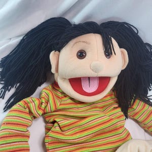 May include: A plush puppet with a smiling face, brown eyes, and long black hair. The puppet wears a long-sleeved shirt with horizontal stripes in green, orange, and red. The mouth is open, revealing a red interior and a pink tongue.