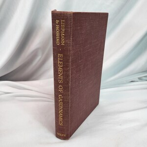 May include: A dark brown hardcover book titled "Elements of Gasdynamics" by Liepmann & Roshko. The spine features gold lettering and the Wiley logo. The book is likely a textbook.