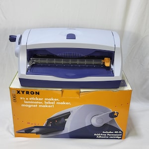May include: A white and purple Xyron 900 sticker maker machine with a manual crank handle on the side. The machine sits on its original orange and white cardboard box, which features product text and an illustration of the device.