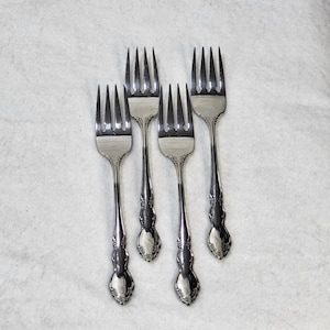 Puede incluir: Cuatro tenedores de metal plateado dispuestos en un patrón vertical. Cada tenedor presenta cuatro dientes y un diseño floral en relieve en el mango. El metal tiene un acabado pulido y reflectante, y los mangos se estrechan hacia la base.