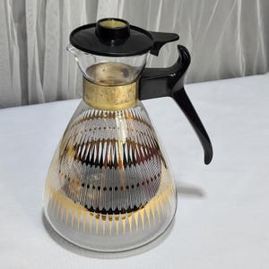May include: Vintage glass pitcher with a black handle and lid. The clear glass body features a gold and white geometric design. A gold band encircles the pitcher's neck. The pitcher is designed for serving beverages.