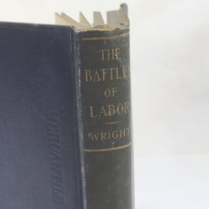 May include: A dark blue hardcover book titled The Battles of Labor by Wright, featuring gold lettering on the spine. The book shows signs of wear along the top edge of the spine and includes the word Mercantile embossed on the front cover.