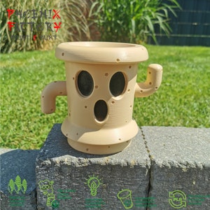 May include: A beige 3D printed planter shaped like a cactus with a face. The planter has a round base and a cylindrical body with a small arm extending out to the side. The face has two black eyes, a black mouth, and a textured surface.