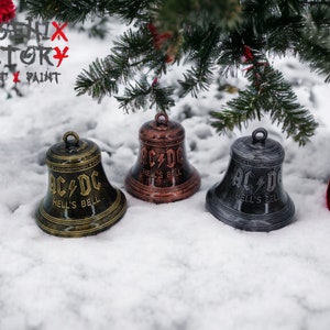 May include: Three metal bells with the AC/DC logo and the words "Hell's Bell" on them. The bells are in different colours: gold, copper, and silver. The bells are sitting on a snowy surface.