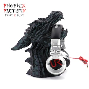 Può includere: Un supporto per cuffie a forma di drago nero che tiene cuffie argento e nere con un cavo rosso. Le cuffie hanno le lettere "UR" in rosso sui padiglioni. Il supporto ha il testo "PHOENIX FACTORY PRINT & PAINT" in rosso e bianco.
