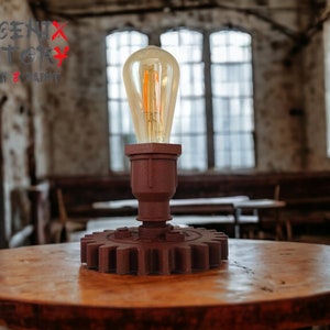 Op de afbeelding: Een rustieke steampunk-stijl tafellamp met een grote tandwielbasis en een heldere glazen lamp. De lamp is aan en geeft een warm gloed. De lamp staat op een houten tafel in een rustieke omgeving.