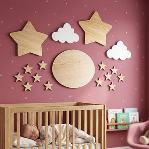 Op de afbeelding: Een set houten wanddecoraties met twee grote sterren, een grote ronde maan, twee witte wolken en tien kleinere sterren. De stukken zijn gerangschikt op een oudroze muur met witte stippen boven een wieg van licht hout met een slapende baby.