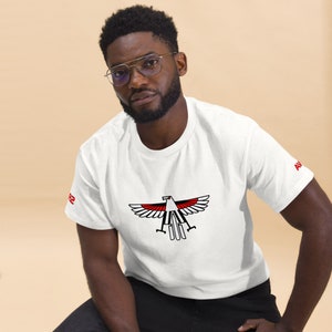 May include: White t-shirt featuring a stylized eagle design in black, red, and white. The eagle graphic is centered on the chest. The letters "AV" are printed in red on the right sleeve, and the number "2" is printed in red on the left sleeve.