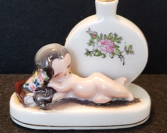 May include: A porcelain figurine of a nude woman lying down with her head resting on a book. She is holding a bouquet of flowers. Behind her is a round porcelain bottle with a floral design. The bottle has a cork stopper.