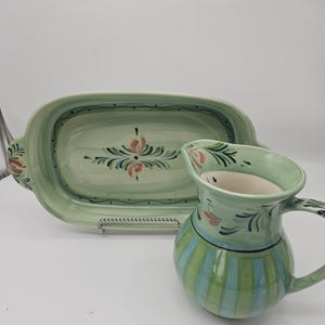 May include: A light green ceramic pitcher and a matching oval serving dish. The pitcher features vertical light blue and green stripes with floral accents. The oval dish has a central hand-painted floral motif and a green border with dots.