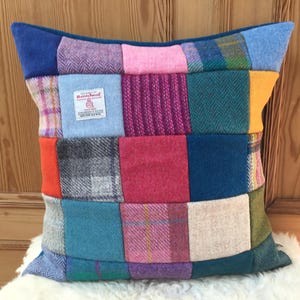 Op de afbeelding: Een patchwork kussen met een verscheidenheid aan kleurrijke wollen stoffen. Het kussen heeft vierkanten in roze, blauw, groen en oranje, met een blauwe rand. Een klein label is zichtbaar op de voorkant. Het kussen ligt op een witte, zachte ondergrond.