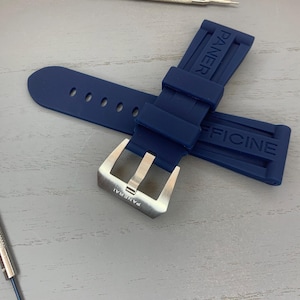 May include: A navy blue watch strap with the brand name "PANERAI" and "OFFICINE" embossed on the rubber. The strap features a silver-colored buckle and several adjustment holes. A watch repair tool is visible in the background.