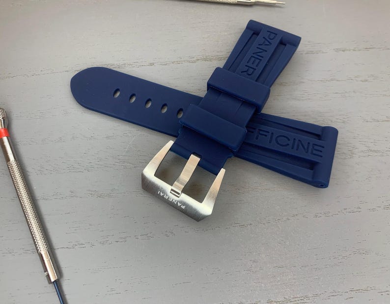 May include: A navy blue watch strap with the brand name "PANERAI" and "OFFICINE" embossed on the rubber. The strap features a silver-colored buckle and several adjustment holes. A watch repair tool is visible in the background.