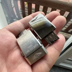 May include: Two silver-toned watch clasps. Each clasp is rectangular with the word "BREITLING 1884" engraved on the surface. The Breitling logo, a stylized bird, is also visible. The clasps are held in a hand.