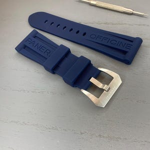 May include: A navy blue watch strap with the brand name "PANERAI OFFICINE" embossed on the rubber. The strap has a silver-colored buckle and is accompanied by two small watch repair tools.