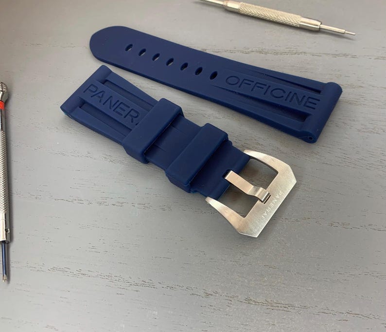May include: A navy blue watch strap with the brand name "PANERAI OFFICINE" embossed on the rubber. The strap has a silver-colored buckle and is accompanied by two small watch repair tools.