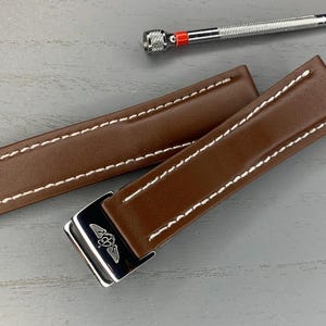 Può includere: Due cinturini per orologio in pelle marrone con cuciture bianche e fibbia argentata. È presente anche un piccolo cacciavite argentato con un accento rosso. I cinturini sono rettangolari e hanno un design cucito.