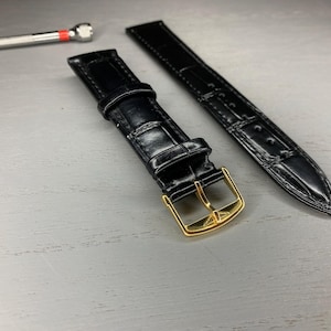 Puede incluir: Correa de reloj de cuero negro con hebilla dorada. La correa es de material texturizado y está sobre una superficie gris claro. Una pequeña herramienta plateada con punta roja está en el fondo.