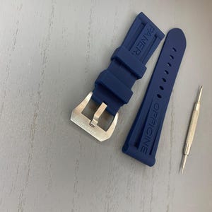 May include: A navy blue watch strap with a silver buckle and the word "PANERAI" embossed on the strap. Two small silver tools are also present.