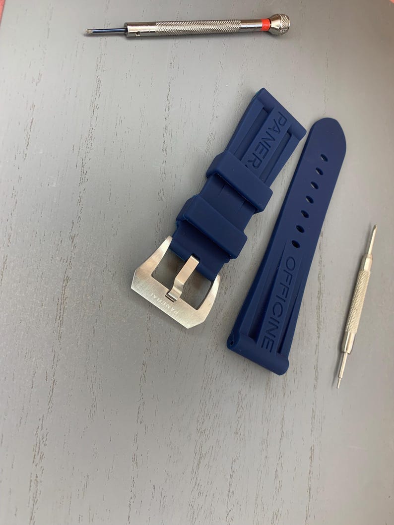 May include: A navy blue watch strap with a silver buckle and the word "PANERAI" embossed on the strap. Two small silver tools are also present.