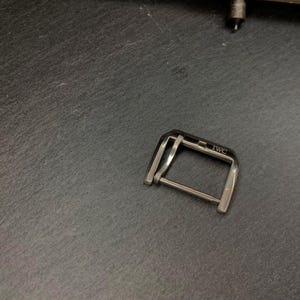 May include: A silver watch buckle with the engraved text "IWC" on a dark grey surface. The buckle is rectangular with a bar in the middle. The watch buckle is a watch accessory.