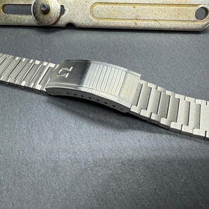 Puede incluir: Una pulsera de reloj de metal plateado con un cierre que tiene el logotipo de Omega.