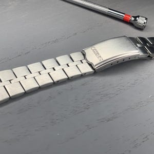 Puede incluir: Una correa de reloj plateada con un acabado de metal cepillado. La correa está compuesta por eslabones rectangulares y presenta un cierre con el nombre de la marca "SEIKO" grabado. Una pequeña herramienta con un acento rojo está en el fondo.