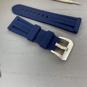 May include: A navy blue watch strap with the word "PANERAI OFFICINE" embossed on the strap. The strap has a silver buckle and is accompanied by two small watch tools. The strap is made of a flexible material.