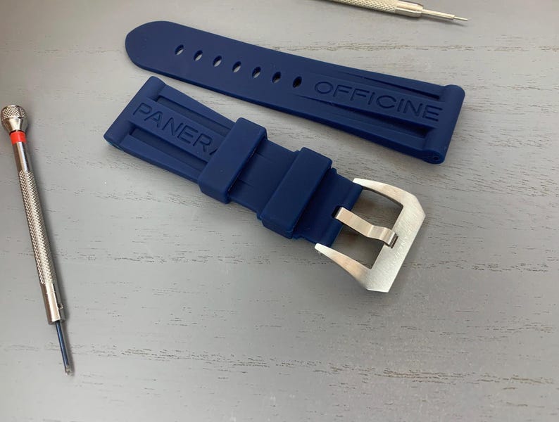 May include: A navy blue watch strap with the word "PANERAI OFFICINE" embossed on the strap. The strap has a silver buckle and is accompanied by two small watch tools. The strap is made of a flexible material.