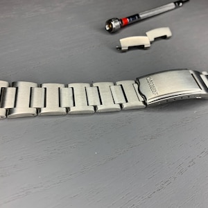 May include: A silver-toned watch band with a clasp, along with a watch tool and a small metal piece. The watch band is made of rectangular links. The clasp is engraved with the word "SEIKO".