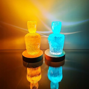 May include: Two decorative glass sculptures, one illuminated in yellow and the other in blue, sit on wooden bases. The textured glass reflects the light, creating a glowing effect. The sculptures are displayed on a reflective surface.