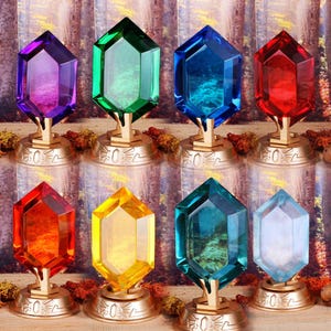 Op de afbeelding: Acht verschillende gekleurde, achthoekige, kristal-achtige edelstenen op gouden standaards. De edelstenen zijn paars, groen, blauw, rood, oranje, geel, turquoise en wit.