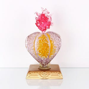 May include: A decorative glass heart-shaped object with a flame-like top. The heart is clear with yellow and pink accents, and sits on a brown and gold base. The flame is a vibrant pink color.