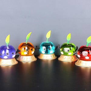 May include: Five colourful, transparent, toy figurines with green stems and black eyes. Each figurine is sitting on a gold base.
