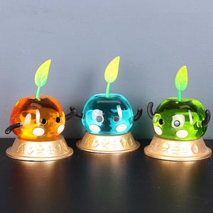 May include: Three colorful, cartoon-like apple figurines. Each has a round, transparent body in orange, blue, and green, with white spots and black arms. They sit on gold-colored bases and have yellow leaves.