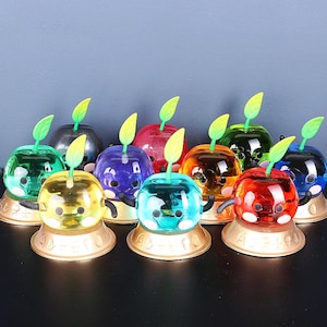 May include: A collection of ten small, colourful, glass apple figurines with green stems and leaves. Each apple has a different colour and a unique facial expression. The apples are arranged in a semi-circle on a white surface.