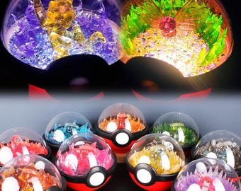 May include: A collection of ten Pokeball-shaped glass ornaments with different colored glitter and figurines inside. The ornaments are arranged in a circle on a white surface.