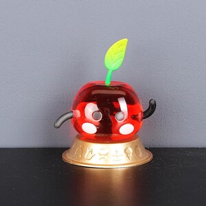 May include: A red cherry-shaped figurine with a green stem and black arms. The figurine is sitting on a gold base with a decorative design.