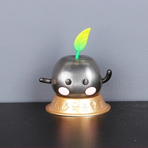 May include: A black and silver figurine with a green leaf on top. The figurine has a round body and two small arms. It is sitting on a gold base with a decorative design.