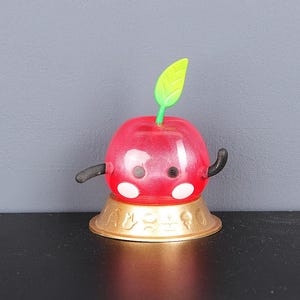 May include: A whimsical red apple-shaped figurine with a green leaf and black curved arms. The apple has white spots and is set on a gold-colored base with hieroglyphic-style symbols. The figurine has a glossy finish.