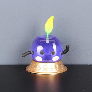 May include: A purple and black character with a green leaf on top, sitting on a gold base. The character has a round body and two small arms.
