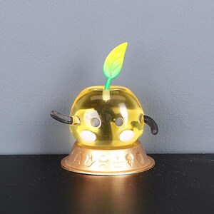 May include: A yellow and green toy with a leaf on top and black arms. The toy is sitting on a gold base.