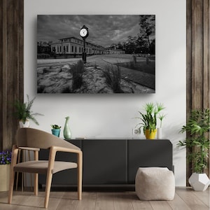 Puede incluir: Fotografía en blanco y negro de un edificio con una torre de reloj. El edificio tiene un diseño simétrico con muchas ventanas. El reloj muestra la hora. La imagen está enmarcada por árboles y un cielo nublado.
