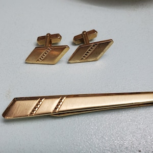 May include: A set of gold-toned cufflinks and a tie bar. The cufflinks are diamond-shaped with a textured design. The tie bar is a long, thin rectangle with a similar textured design.