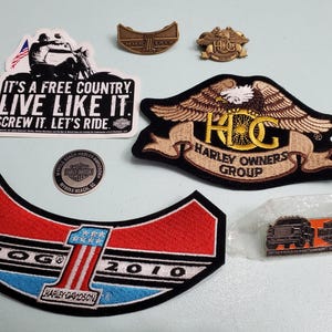 May include: A collection of Harley Davidson patches and pins. The patches are embroidered with the Harley Owners Group logo, a Harley Davidson motorcycle, and the text "It's a free country. Live like it. Screw it. Let's ride.". The pins are metal and feature the Harley Davidson logo and a motorcycle.