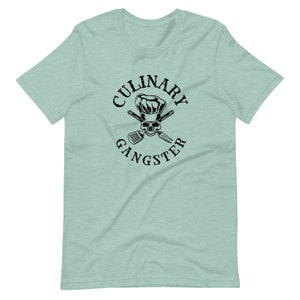 May include: A light blue t-shirt with a black graphic of a skull wearing a chef's hat and crossed kitchen utensils. The text "CULINARY GANGSTER" is written in a circle around the graphic.
