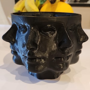 May include: A black, 3D-printed planter with multiple faces around its exterior. The faces have closed eyes and subtle features. The planter is on a white surface. Bananas and a yellow fruit are visible in the background.
