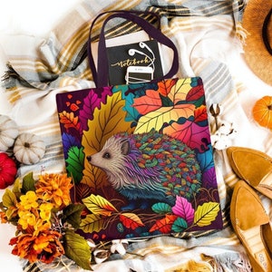 May include: A tote bag with a colorful hedgehog design featuring autumn leaves. The bag is made of canvas and has a black handle. The hedgehog is surrounded by leaves in shades of red, orange, yellow, and green.