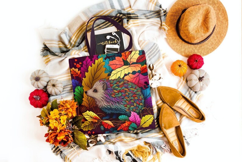 May include: A tote bag with a colorful hedgehog design featuring autumn leaves. The bag is made of canvas and has a black handle. The hedgehog is surrounded by leaves in shades of red, orange, yellow, and green.